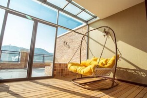 Una habitación con un amplio ventanal que da a un balcón, un sillón colgante con cojines amarillos y una pared de piedra.