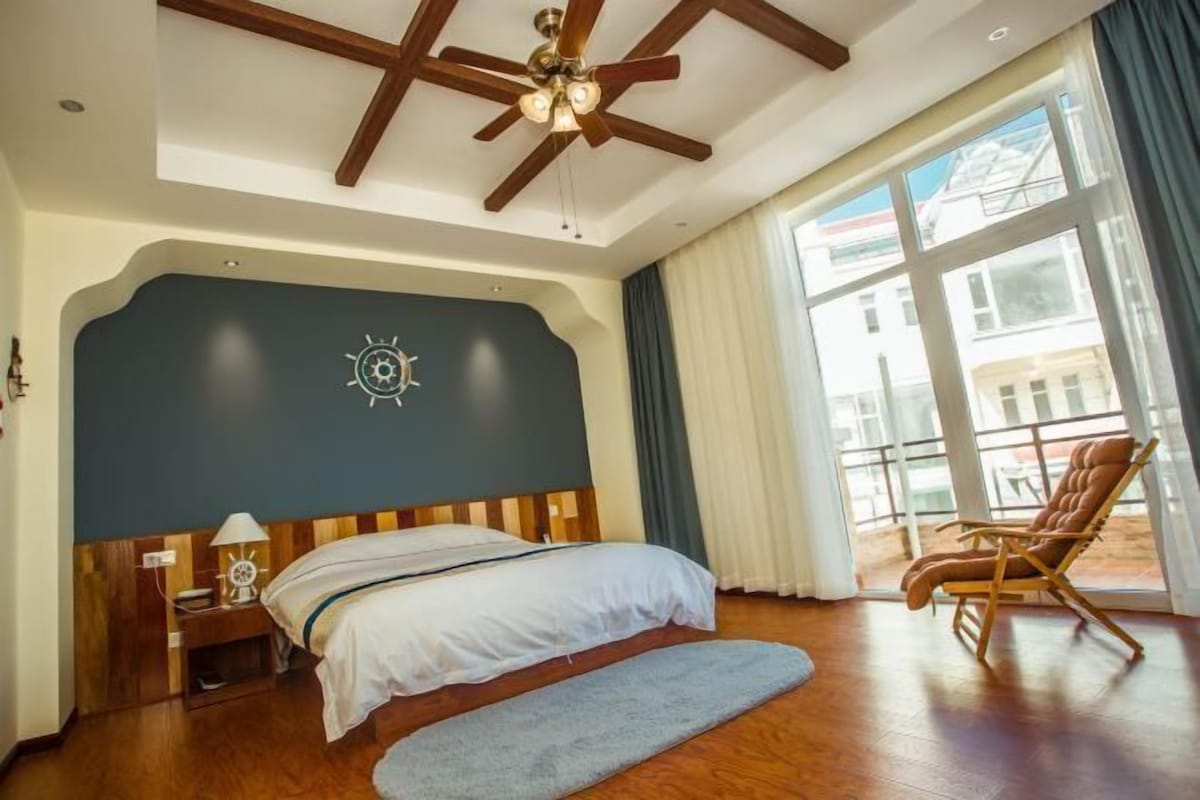 A bedroom with a large bed, a wooden ceiling fan, a nightstand with a lamp, a chair, and a view of the outside through a window.