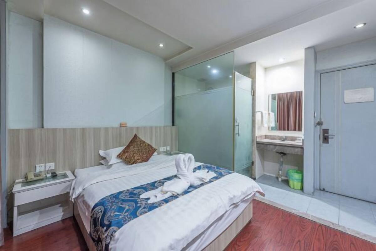 Une chambre d’hôtel comprenant un lit, une table de chevet, une cabine de douche en verre, une salle de bain avec un lavabo et une porte.