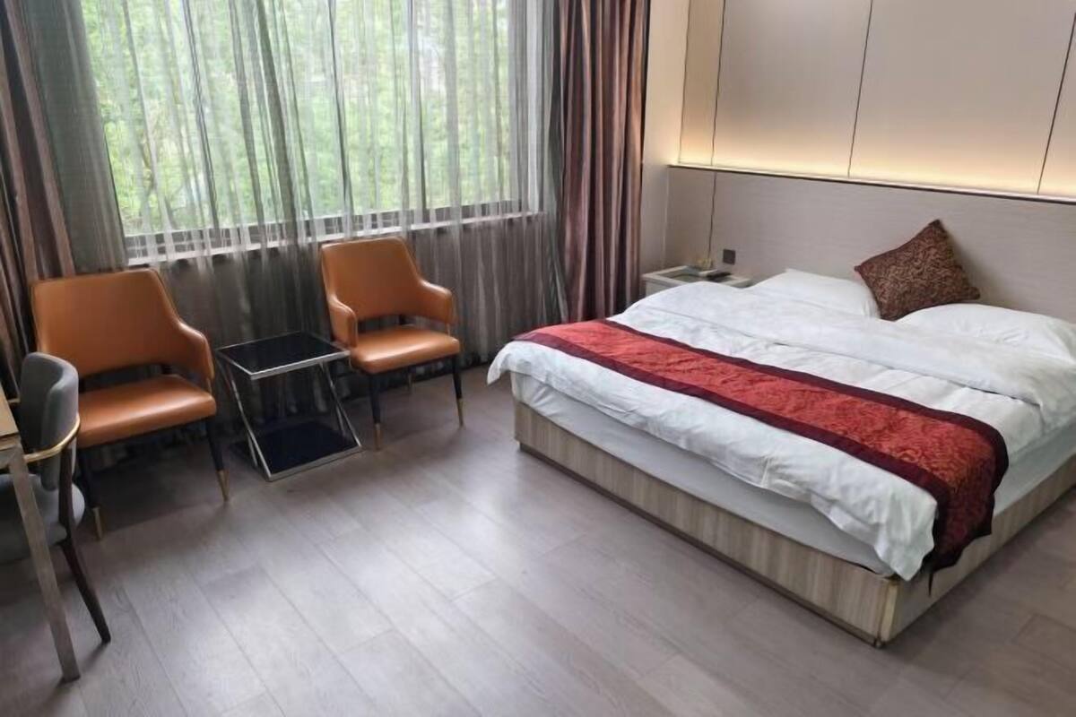 Une chambre d’hôtel avec un lit, deux chaises, une petite table et une fenêtre avec des rideaux.