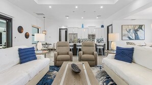 A modern living room with two sofas, a coffee table, and a dining area in the background. - Gorgeous! Heated, Saltwater Pool and Spa, Custom Outdoor Kitchen - Villa Still Waters - Roelens (Cape Coral)