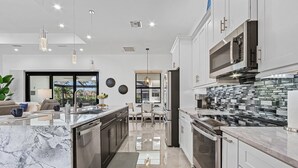 A modern kitchen with a marble countertop, stainless steel appliances, and a view to the outdoor patio. - Gorgeous! Heated, Saltwater Pool and Spa, Custom Outdoor Kitchen - Villa Still Waters - Roelens (Cape Coral)