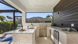 A modern kitchen with a built-in stove, sink, and refrigerator, large windows offering a view of palm trees and a house. - Gorgeous! Heated, Saltwater Pool and Spa, Custom Outdoor Kitchen - Villa Still Waters - Roelens (Cape Coral)