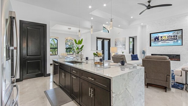 A modern kitchen with dark cabinets, a marble island, and pendant lighting. - Gorgeous! Heated, Saltwater Pool and Spa, Custom Outdoor Kitchen - Villa Still Waters - Roelens (Cape Coral)