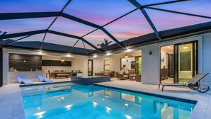 A modern indoor swimming pool with a glass ceiling, lounge chairs, and a well-lit patio area. - Gorgeous! Heated, Saltwater Pool and Spa, Custom Outdoor Kitchen - Villa Still Waters - Roelens (Cape Coral)