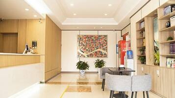 A hotel lobby with a reception desk, a dining area with chairs, a large abstract painting, and a bookshelf with various items.