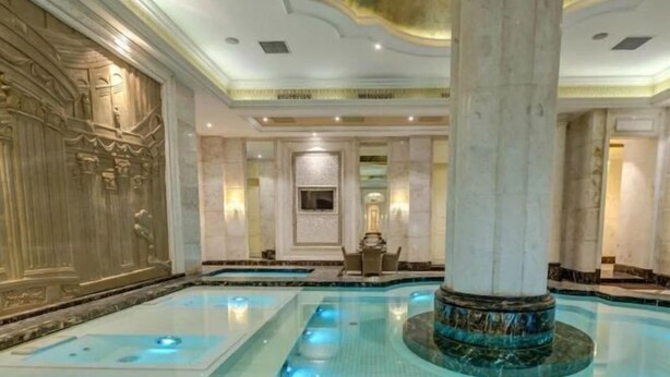 Un área de piscina interior lujosa, con suelo de mármol, una columna central y un diseño de techo imponente.