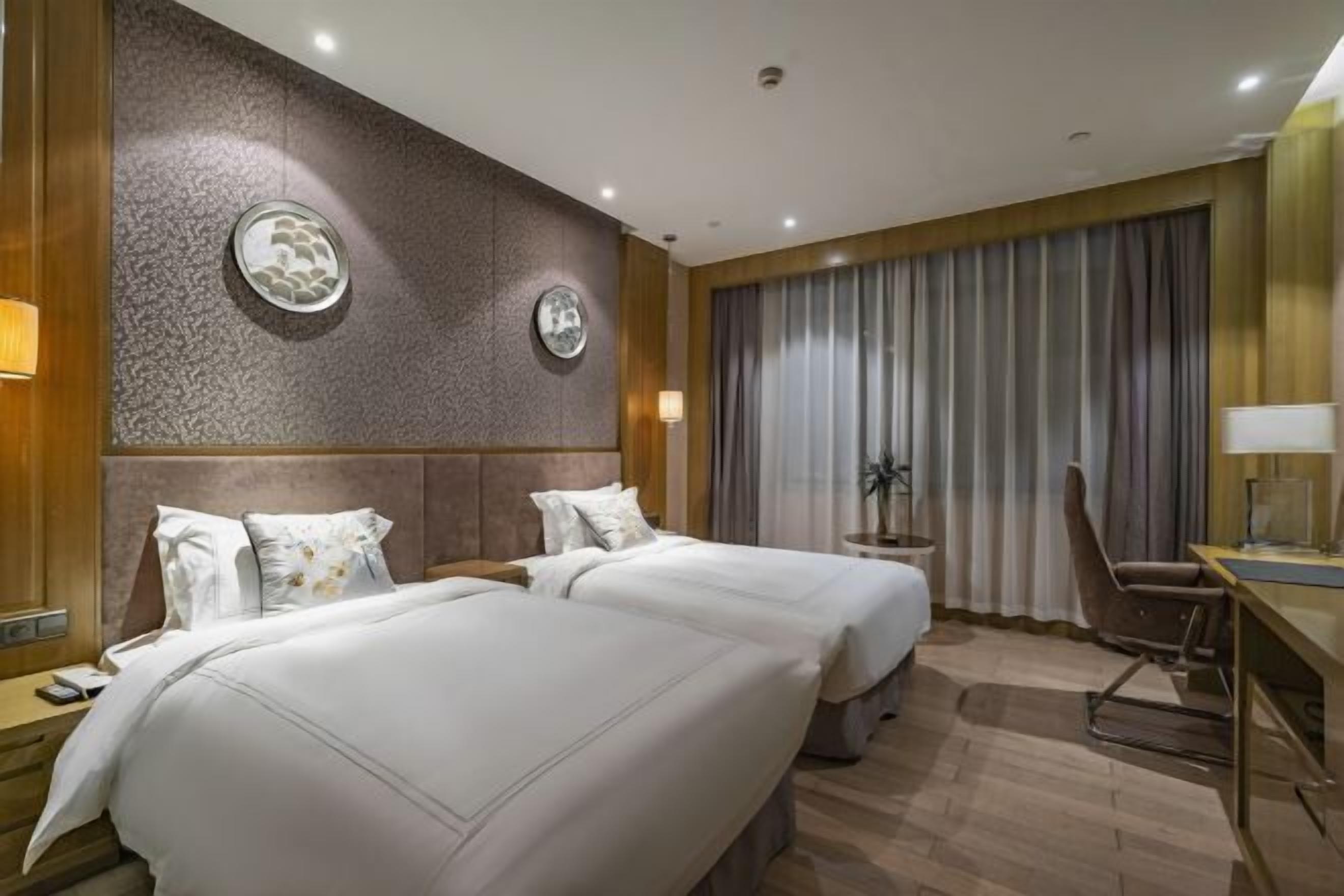 A hotel room with two beds, a desk, a chair, a lamp, and a large window with curtains.