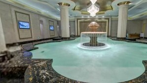 Una amplia piscina interior con una fuente central, rodeada de suelos de mármol y columnas.