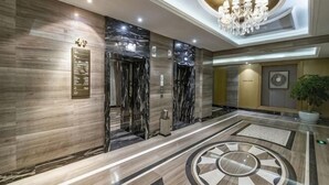 Un vestíbulo de hotel con suelo de mármol, ascensores, una araña y un gran diseño circular en el piso.