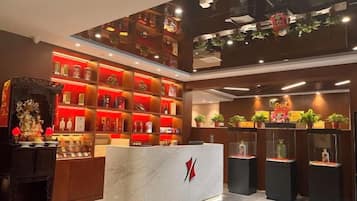 A hotel lobby with a reception desk, shelves with items, and potted plants.