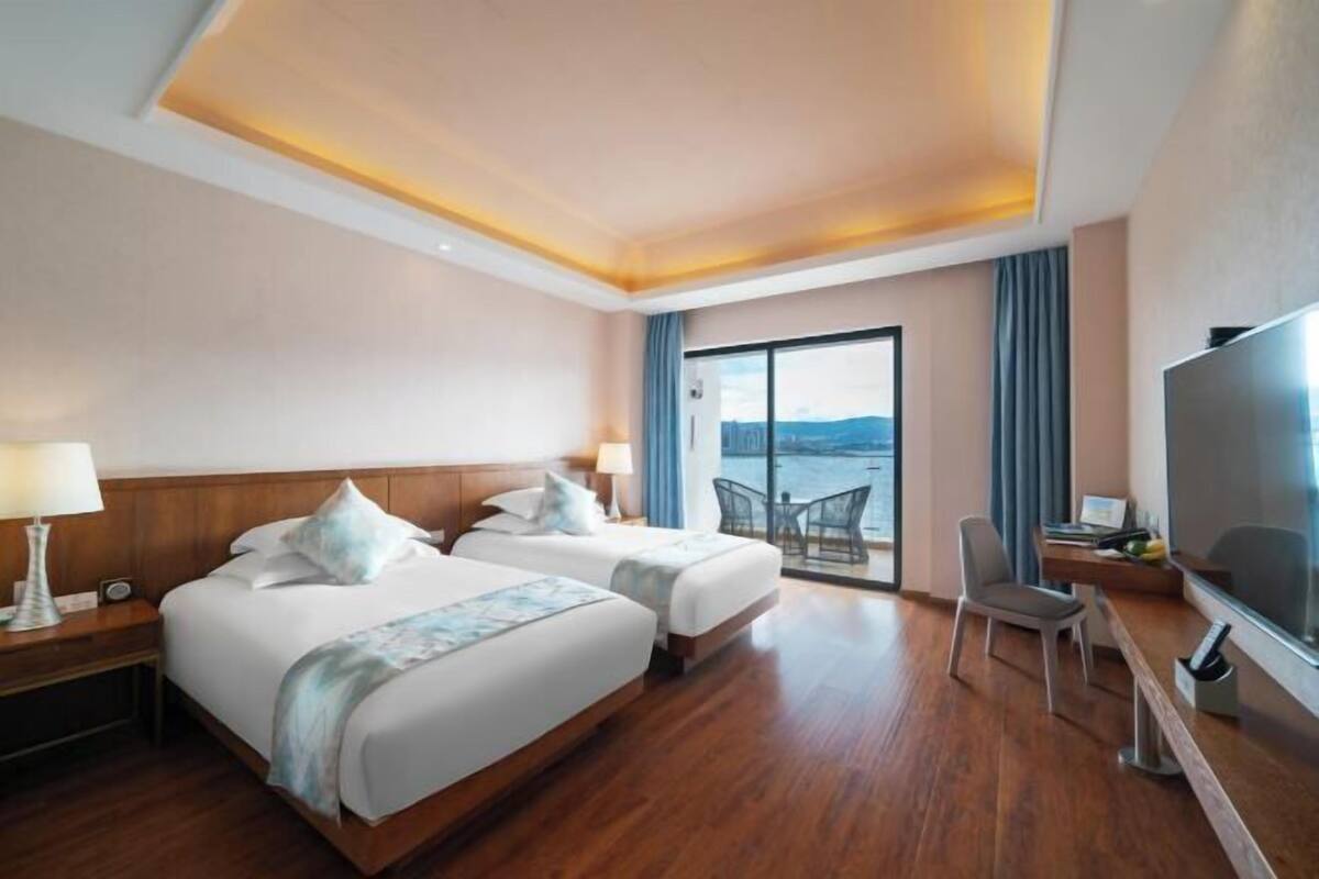 A hotel room with two beds, a desk, a chair, a TV, and a view of the sea.