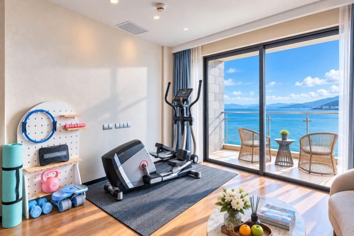 A fitness room with exercise equipment, a yoga mat, and dumbbells. There is a large window with a view of the sea.