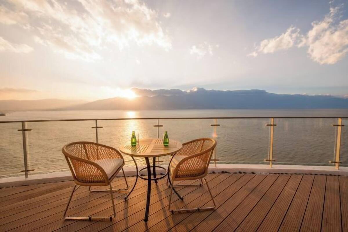A wooden deck with two wicker chairs and a table, overlooking a body of water with mountains in the distance.