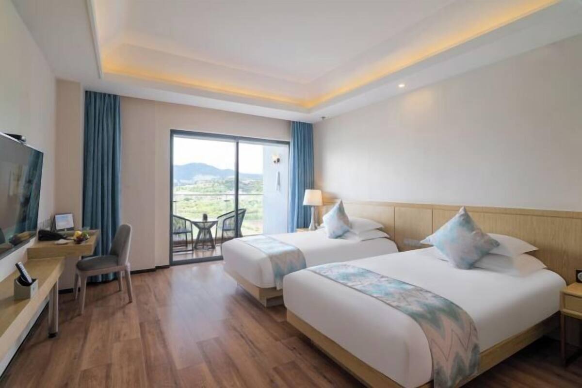 A hotel room with two beds, a desk with a chair, a TV, and a large window offering a view of the outdoors.