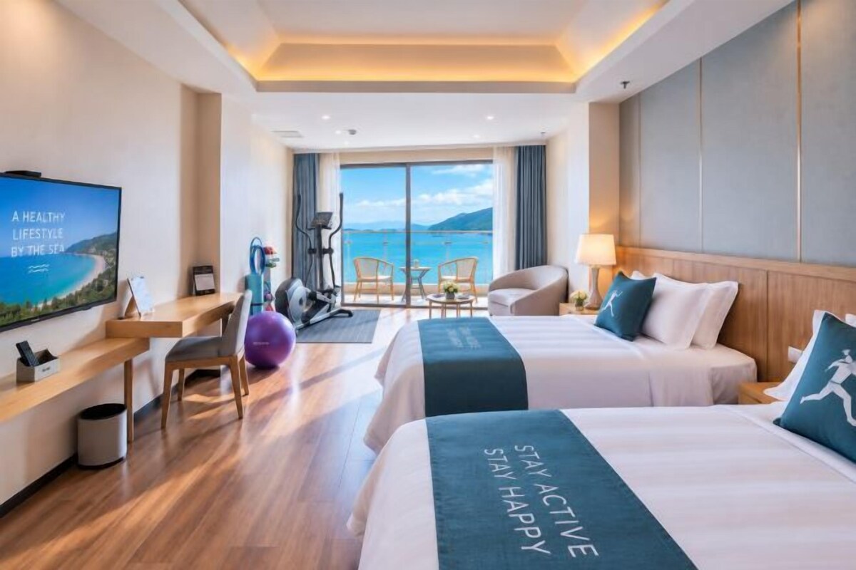 A hotel room with two beds, a desk, a TV, a balcony with a view of the sea, and exercise equipment.