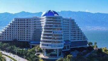A modern hotel with a unique architectural design featuring a central dome and multiple balconies, located near a body of water with mountains in the background.