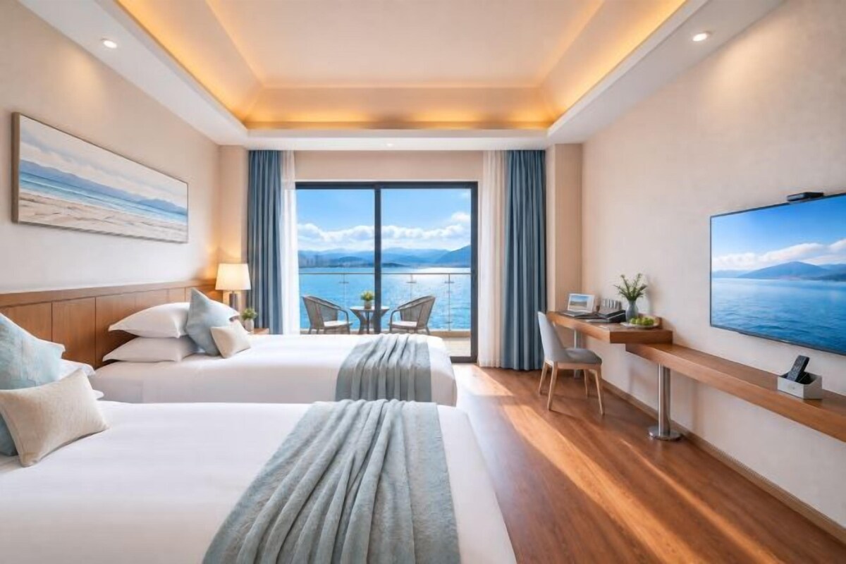 A hotel room with two beds, a desk, a TV, and a balcony with a view of the sea.