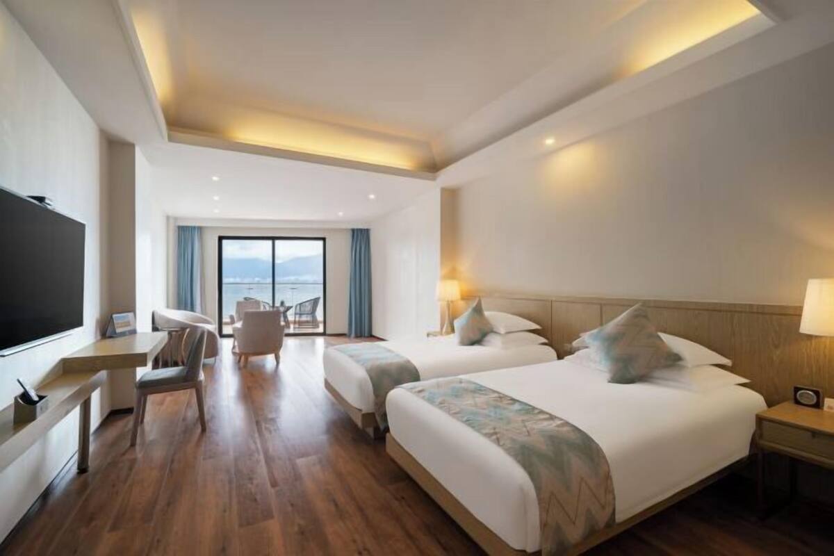 A hotel room with two beds, a desk, a chair, a TV, and a view of the sea.