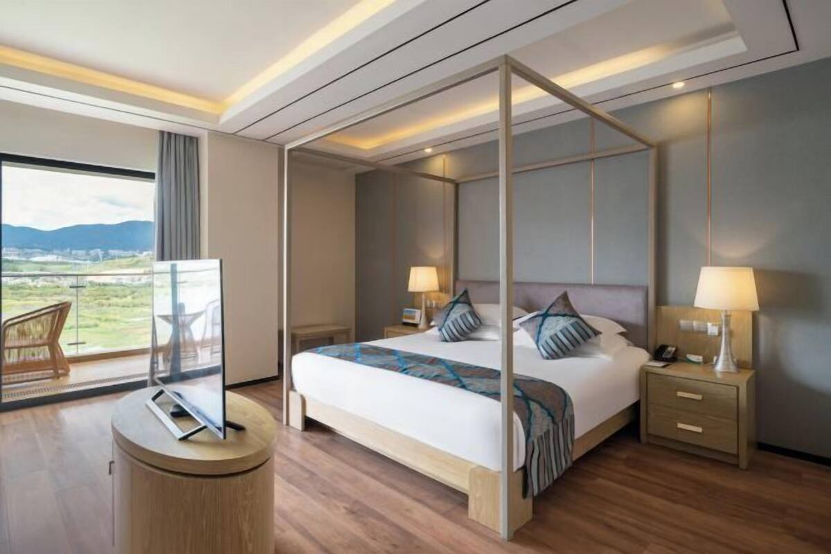 A modern hotel room with a large bed, bedside tables with lamps, a view of the outdoors, and a round wooden side table.