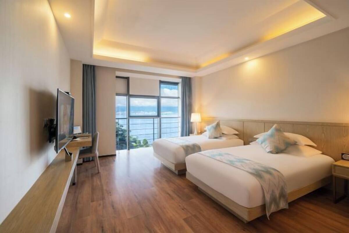 A hotel room with two beds, a TV, a desk, and a large window offering a city view.