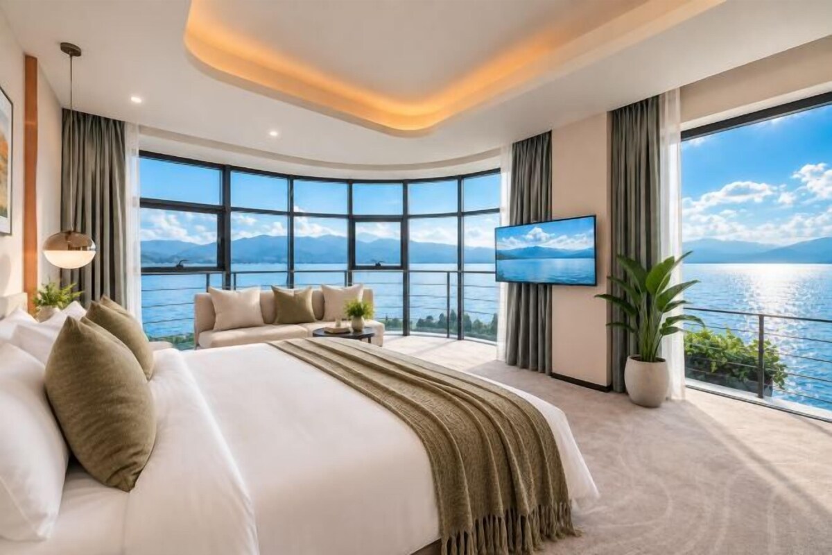 A modern hotel room with a large bed, a sofa, a TV, and a view of the sea.