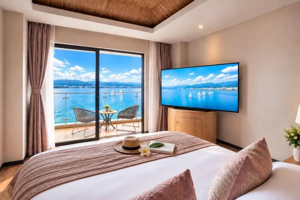 A hotel room with a large bed, a TV, a balcony with a view of the sea, and a dining area with chairs.