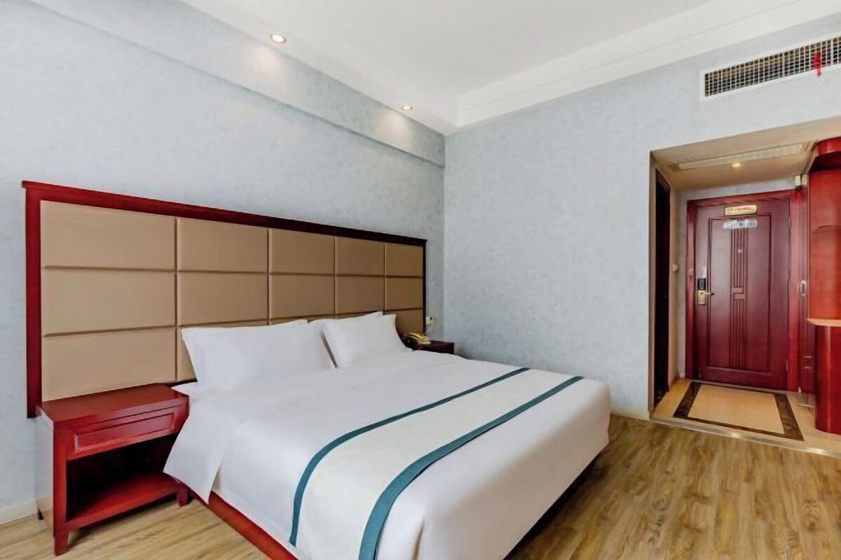 A hotel room with a large bed, a wooden nightstand, a red door, and a light-colored wall.