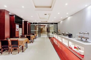 Un comedor de hotel con mesas y sillas, una barra de buffet con exhibiciones de comida, y una vista despejada de la entrada.