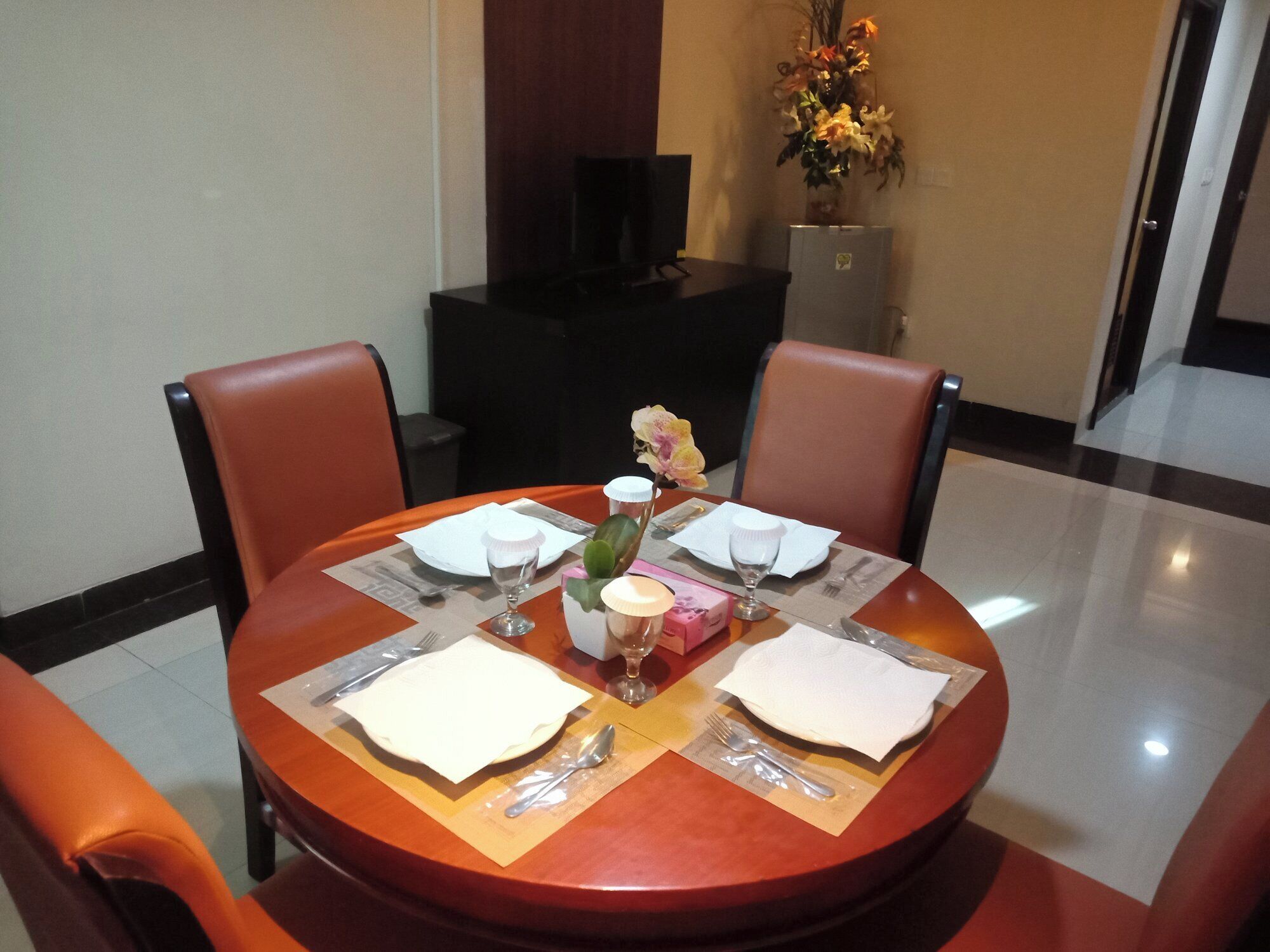 A dining table set for four with a vase of flowers, a television in the background, and a tiled floor.