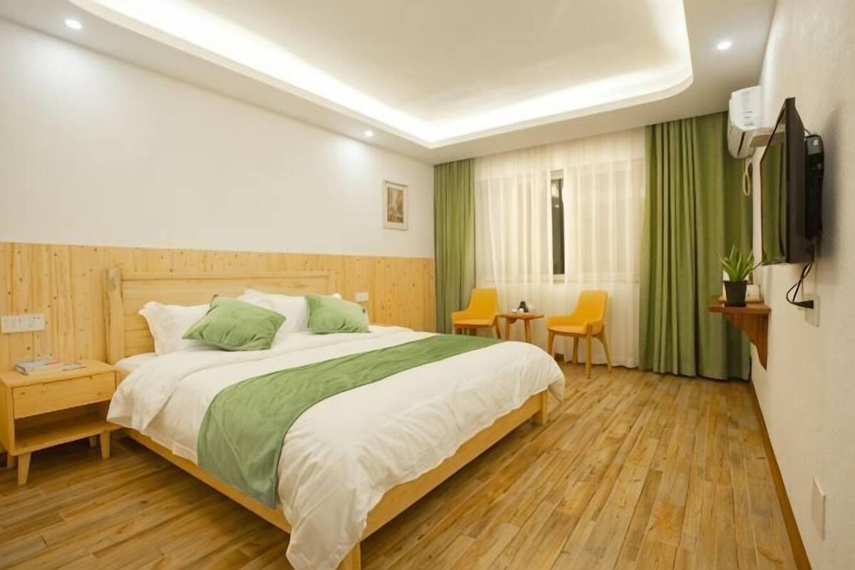 Habitación de hotel con cama de madera, mesita de noche, silla, pequeña mesa, televisor y ventana con cortinas.