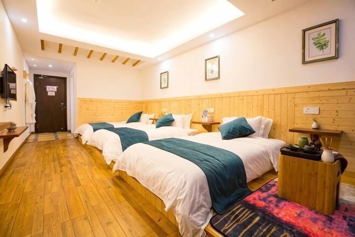 Habitación de hotel con tres camas, piso de madera, cabecera de madera, una mesita con lámpara y un cuadro en la pared.