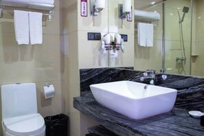 A modern bathroom with a white toilet, a large white sink on a marble countertop, and a glass shower enclosure.