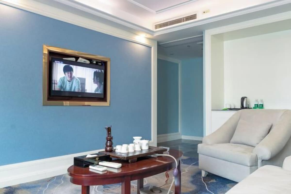 A hotel room with a television mounted on the wall, a small wooden table with cups and a tray, and a beige armchair.