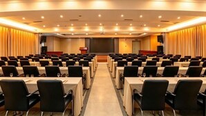 Un salón de conferencias con filas de mesas y sillas, un atril y una pantalla grande al frente.