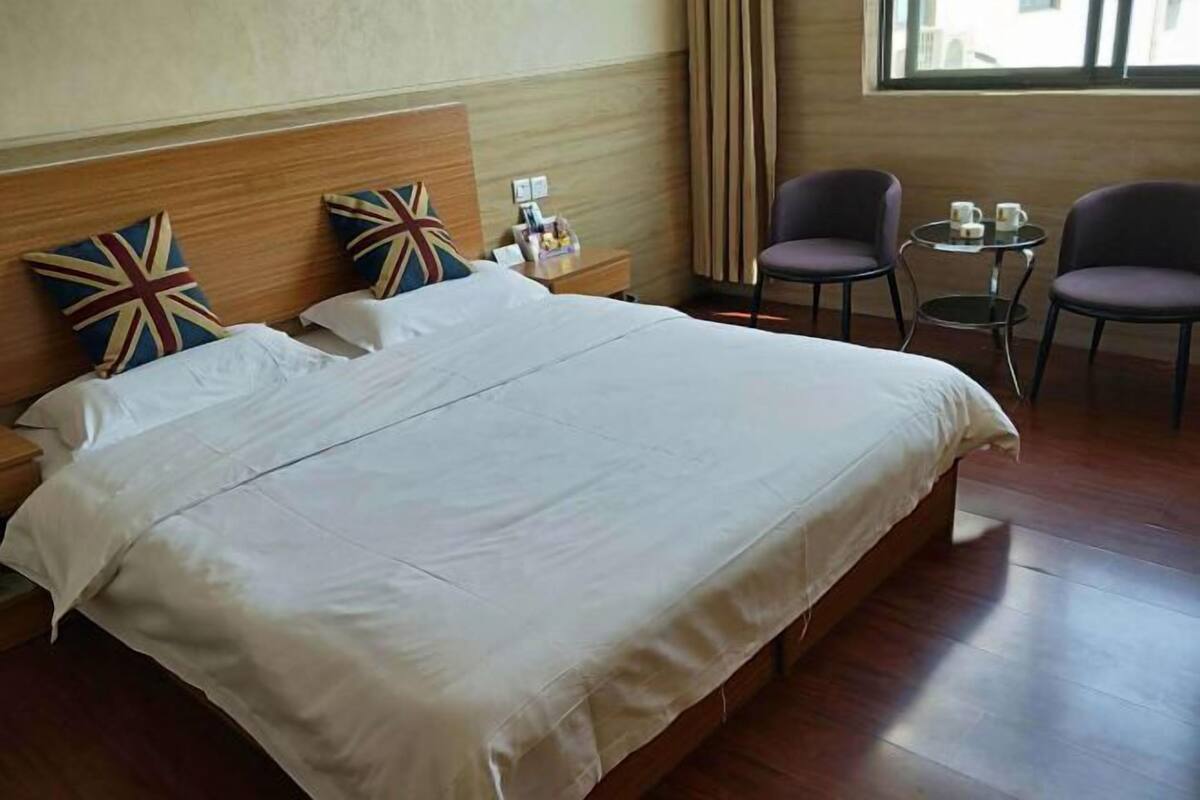 A hotel room with a large bed, two pillows with a Union Jack design, a wooden headboard, a nightstand, two chairs, a small round table with two cups, and a window with curtains.