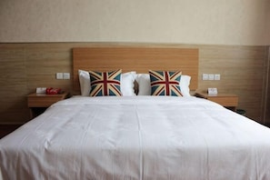 Cama com roupa de cama branca, duas almofadas com estampas da Union Jack, cabeceira de madeira e duas mesas de cabeceira com objetos vermelhos e brancos.