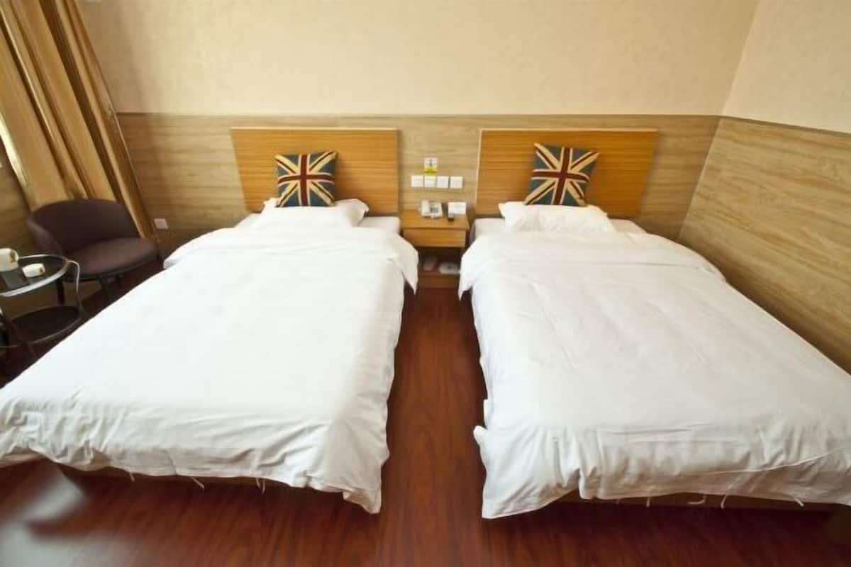 A hotel room with two single beds, a wooden headboard, a small table with a chair, and a nightstand with a lamp.