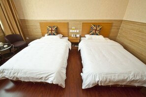 Habitación de hotel con dos camas individuales, cabecero de madera, una mesita pequeña con una silla y una mesita de noche con lámpara.