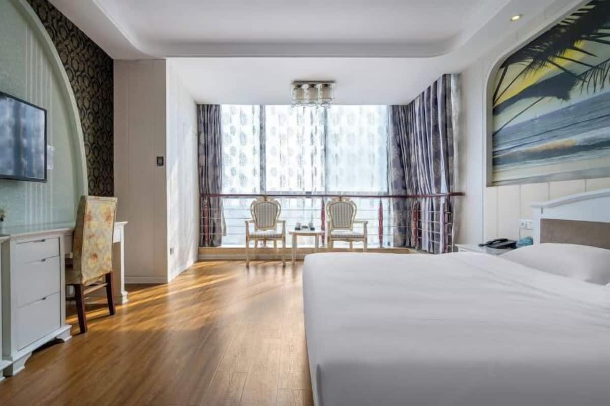 Une chambre d’hôtel avec un grand lit, un bureau avec une chaise, une petite table avec deux chaises, une grande fenêtre avec des rideaux et un tableau au mur.
