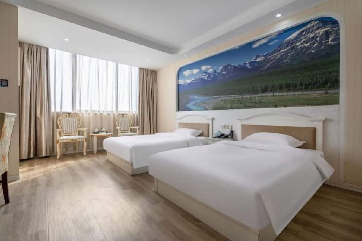 Une chambre d’hôtel avec deux lits, une grande fenêtre avec des rideaux, une chaise et une fresque murale représentant un paysage de montagne.