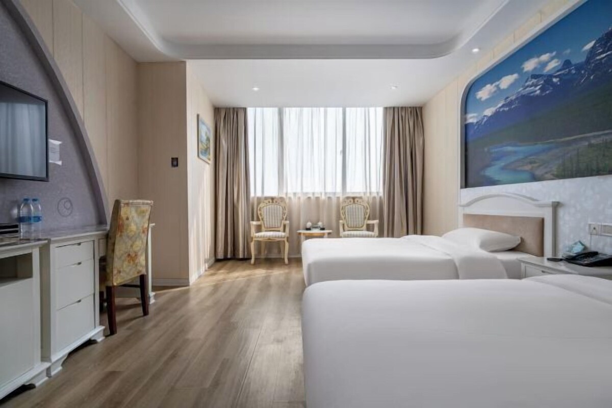 Ein Hotelzimmer mit zwei Betten, einem Schreibtisch mit Stuhl, einem kleinen Tisch und einem Bild an der Wand.