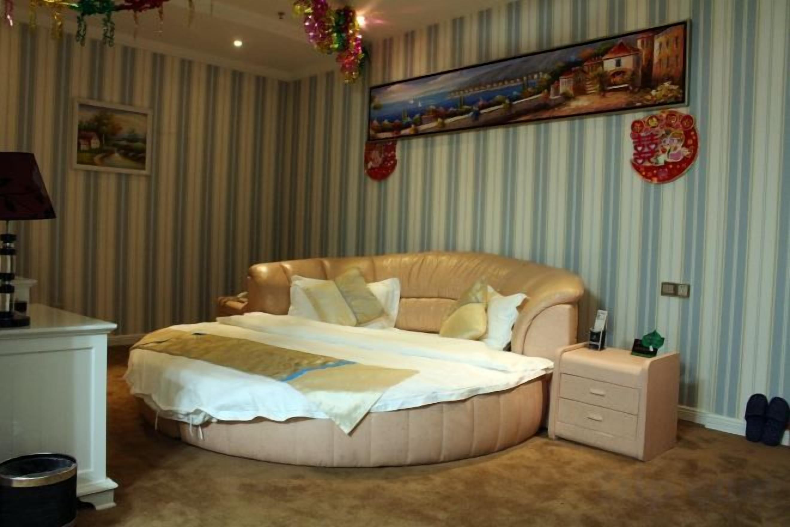 A bedroom with a large round bed, a bedside table, a nightstand, and a wall-mounted painting.