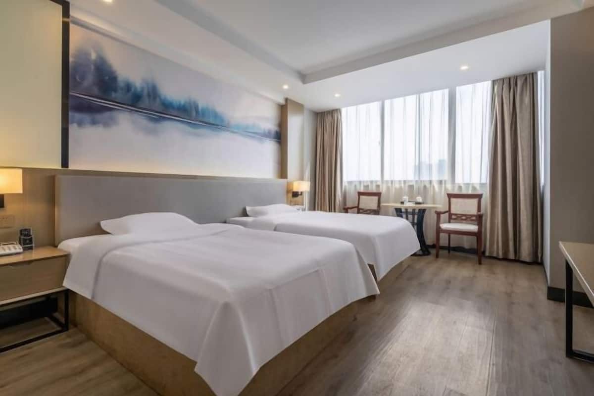 Une chambre d’hôtel avec deux lits, un bureau, une chaise, une grande fenêtre avec des rideaux et un tableau au mur.