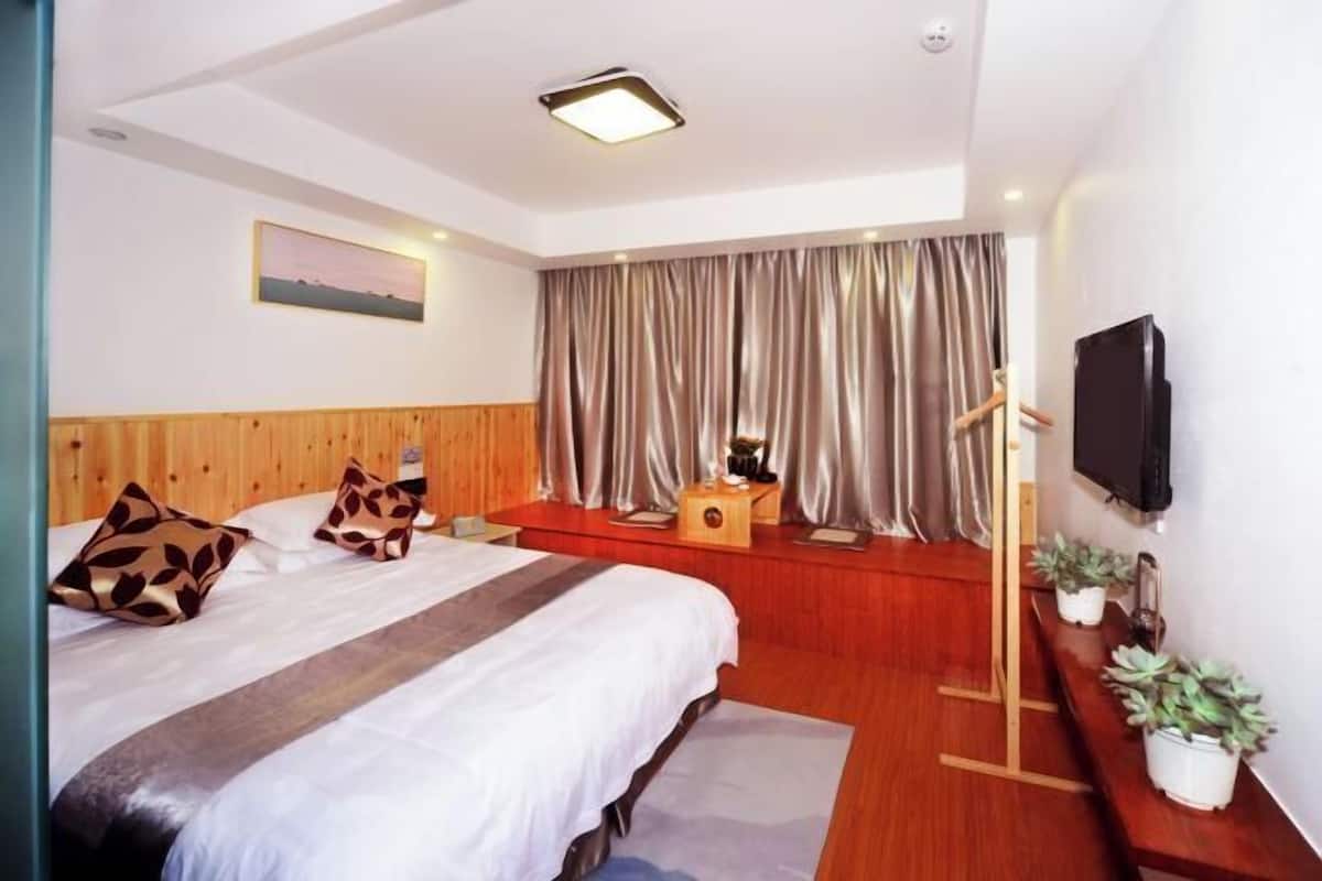 A modern hotel room with a wooden headboard, a large bed with patterned pillows, a bedside table with a lamp, a TV mounted on the wall, and a window with curtains.
