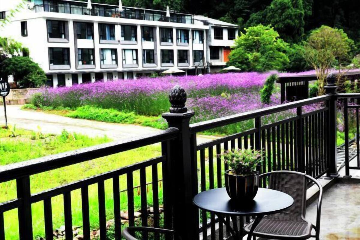 A balcony with a black railing, a small round table with a potted plant, and a view of a building with multiple windows and a purple flower garden.
