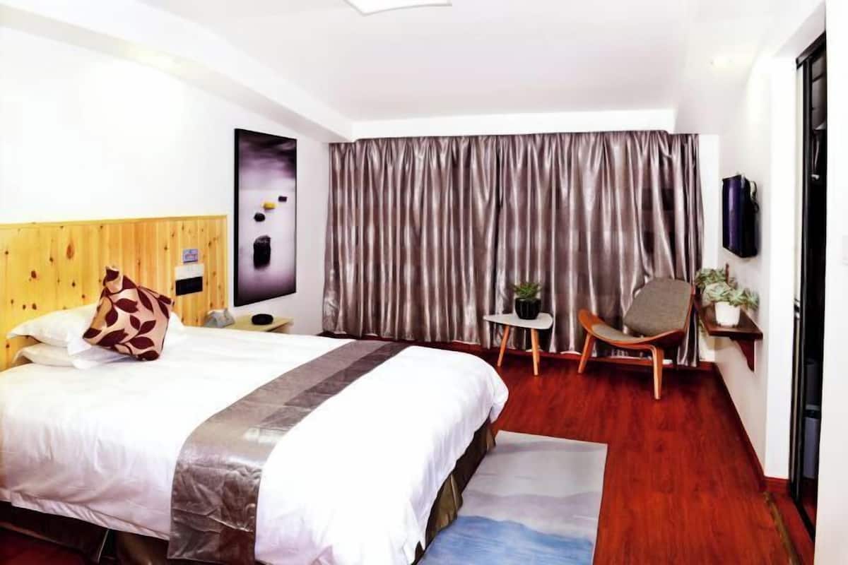 A hotel room with a bed, a small table with a plant, a chair, and a large window with curtains.