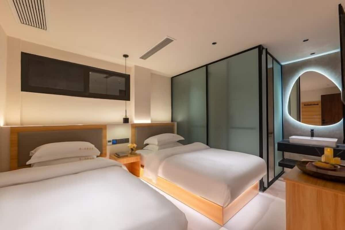 A modern hotel room with two beds, a bedside table, a mirror, and a bathroom with a sink and mirror.