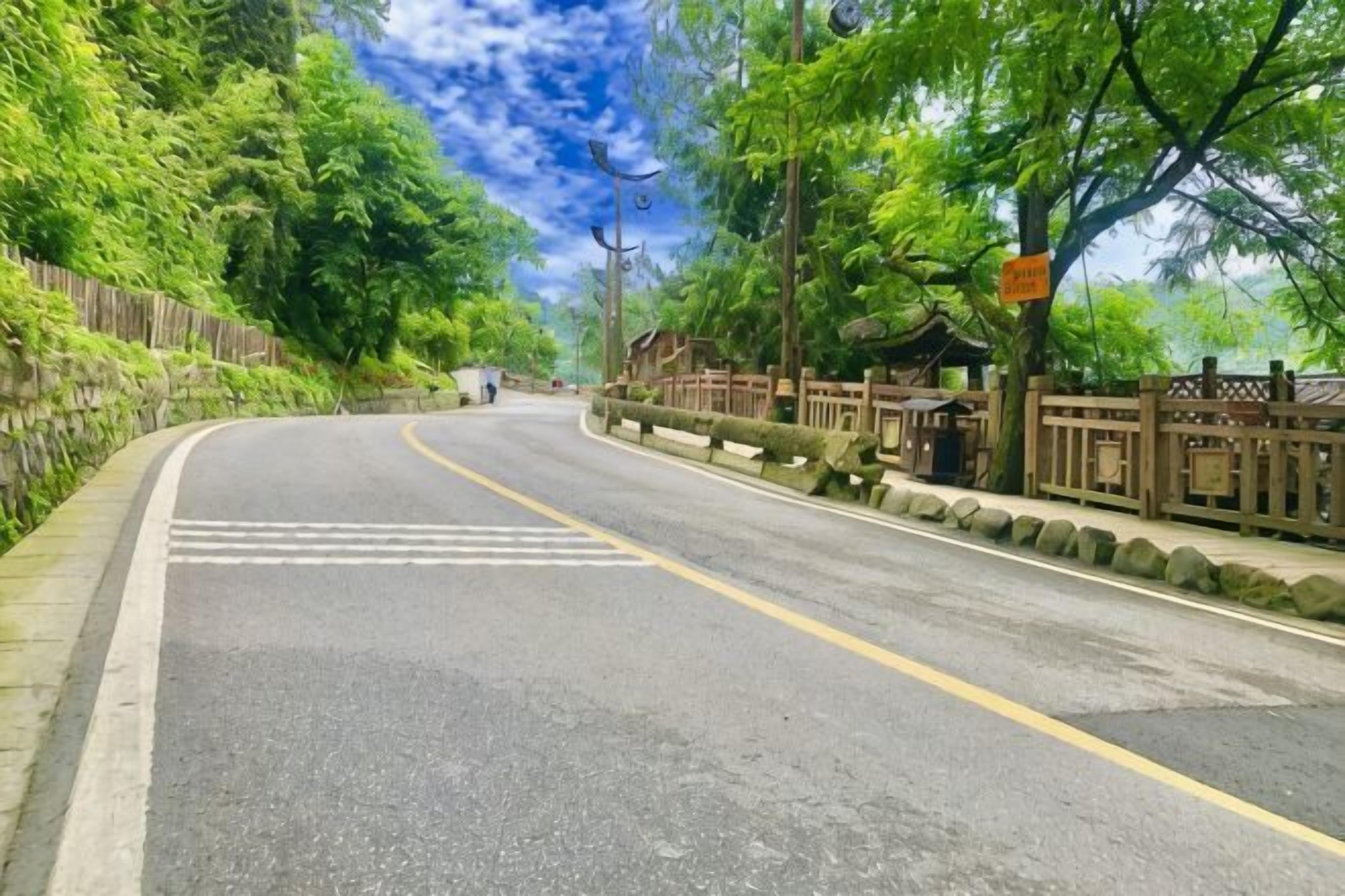 A curved road with a yellow dividing line, surrounded by greenery and a wooden fence.