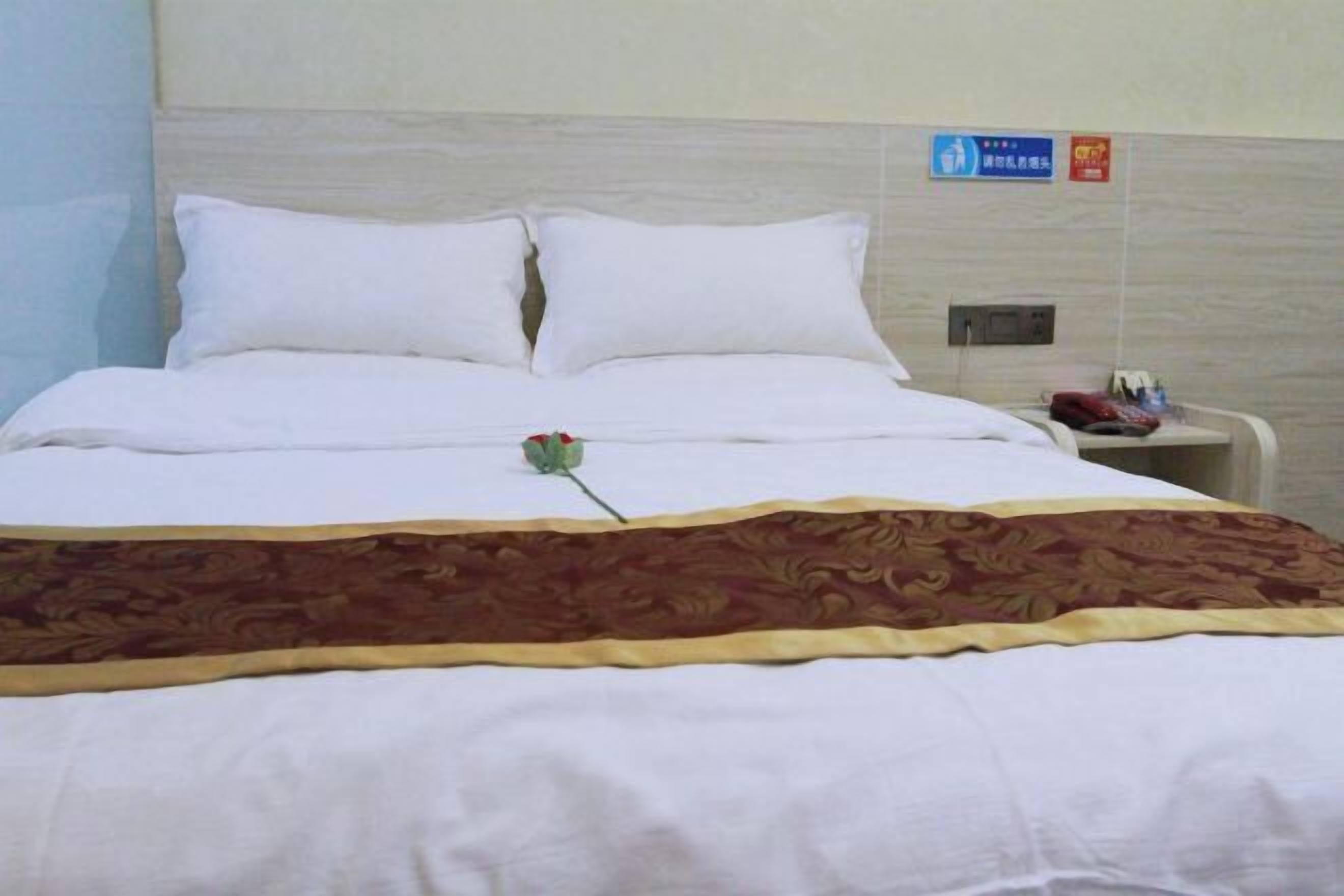 A hotel room with a neatly made bed, a bedside table with a phone and a bottle, and a sign on the wall.